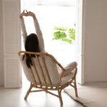 A woman stretches while sitting in a chair facing a window.