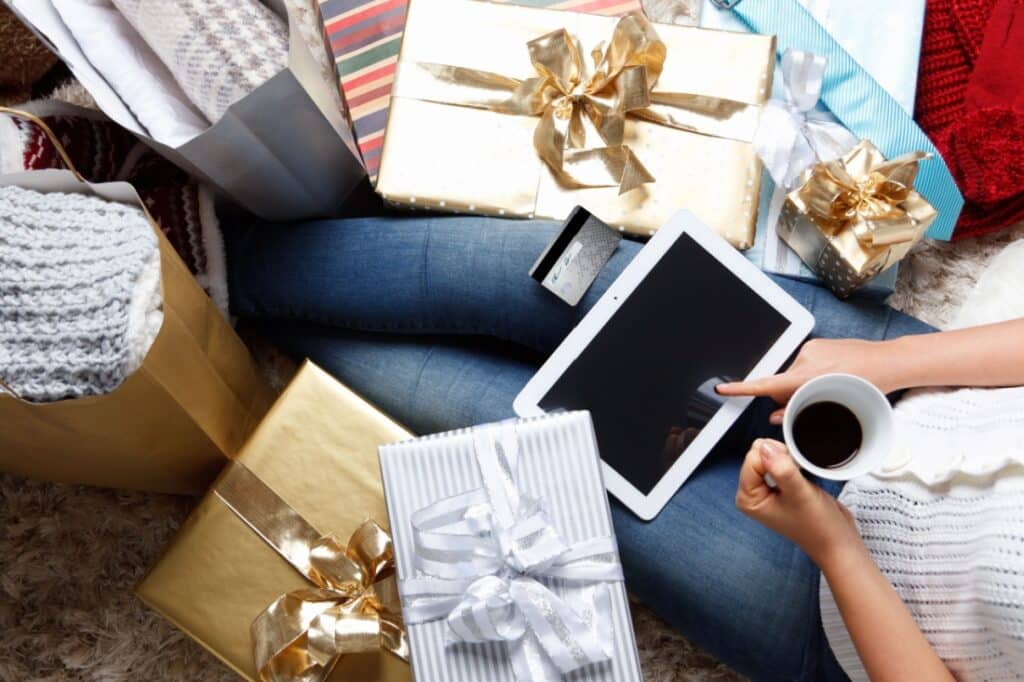 A person holds a coffee mug and taps on their iPad while surrounded by gifts.