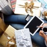 A person holds a coffee mug and taps on their iPad while surrounded by gifts.