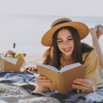 young happy traveler woman in straw hat reading book at beach