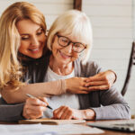 An adult daughter helps a senior woman complete a form at home.