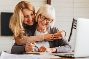 An adult daughter helps a senior woman complete a form at home.