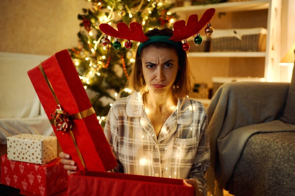 A woman with a festive reindeer headband looks disappointed as she opens a gift.