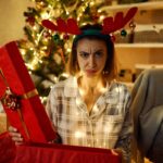 A woman with a festive reindeer headband looks disappointed as she opens a gift.