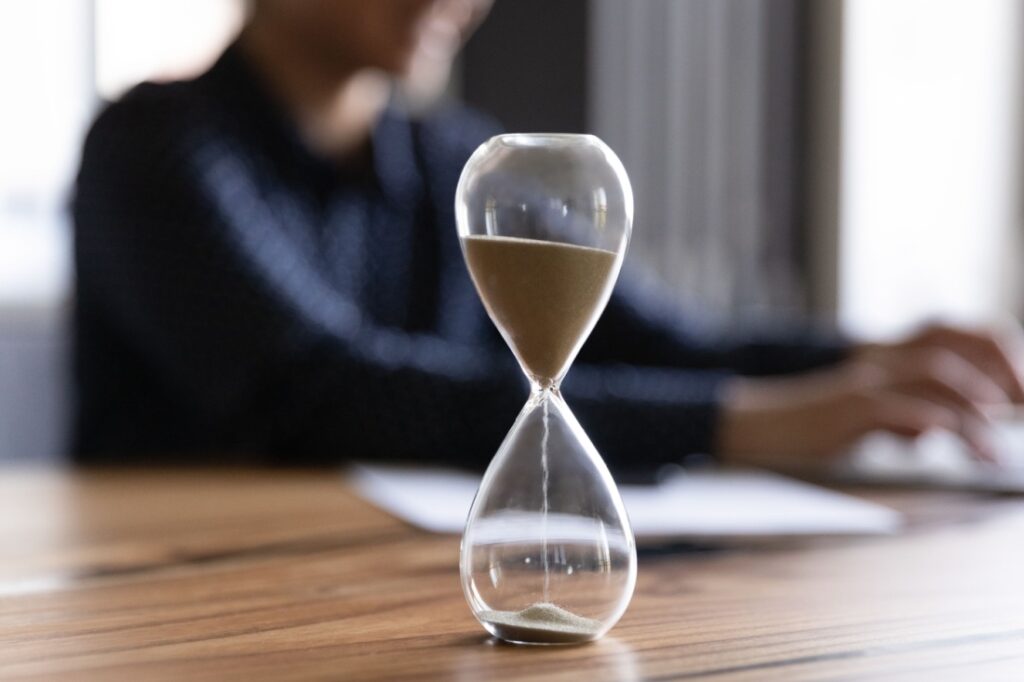 A close up of an hourglass. In the background, someone types on their computer.