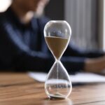 A close up of an hourglass. In the background, someone types on their computer.