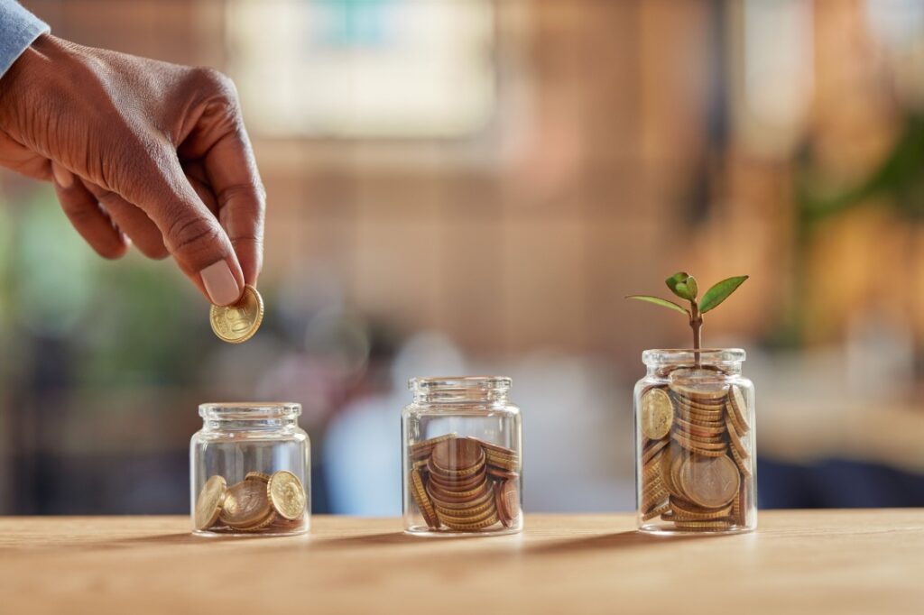 A hand deposits money into a jar. There are two jars with more money, and a plant is growing from the one with the most.