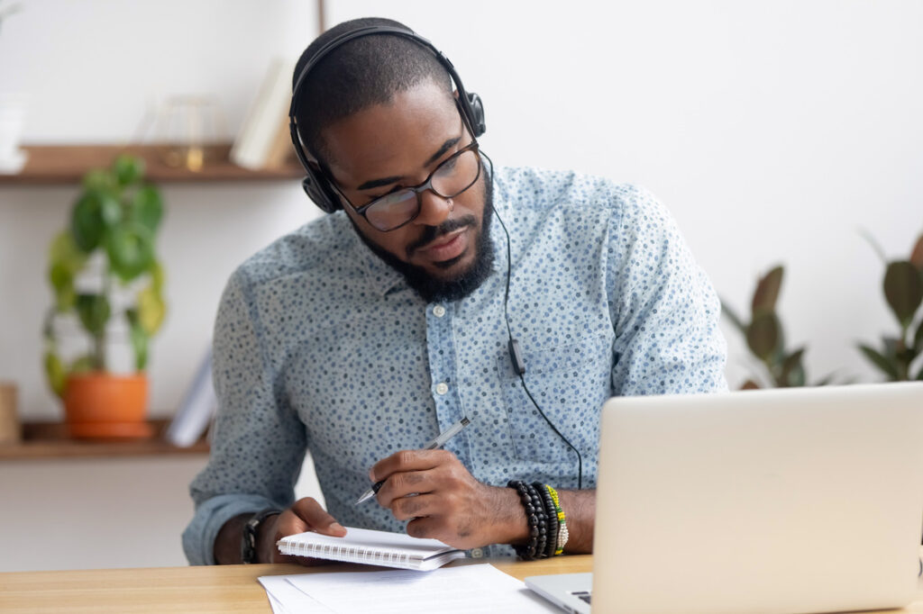 a man taking notes on a notepad while looking at a laptop and wearing headphones