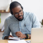 a man taking notes on a notepad while looking at a laptop and wearing headphones