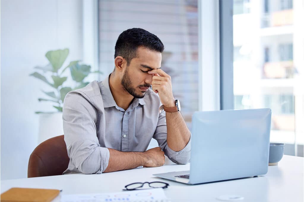 A stressed out man sitting at a desk with a laptop, pinching the bridge of his nose