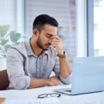 A stressed out man sitting at a desk with a laptop, pinching the bridge of his nose