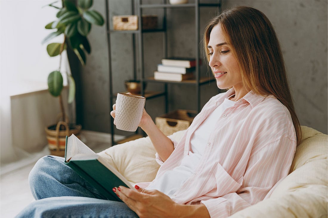 Side view young woman sits in armchair reading book drinking coffee