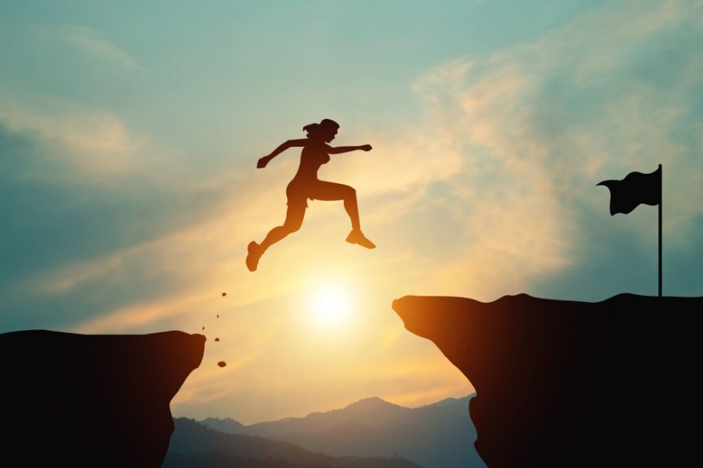 Silhouette of a woman jumping across as gap between two cliffs to reach a finish line flag.