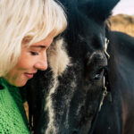 Blonde woman with black horse in outdoor scenic destination hugging