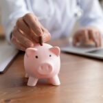 Close up woman putting coin into pink piggy bank, sitting at work desk