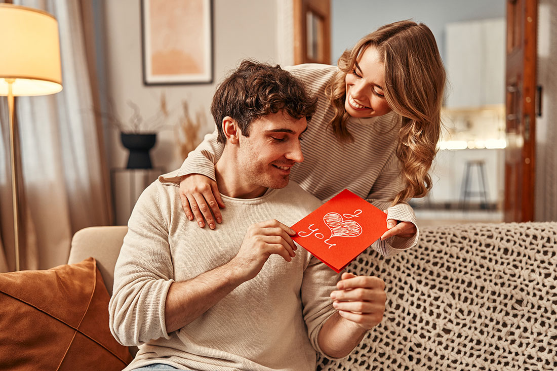 A woman gives a greeting card to her man with the inscription I love you in the living room at home