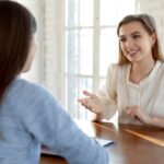 Two women sit across from each other, one is smiling and greeting the other warmly.