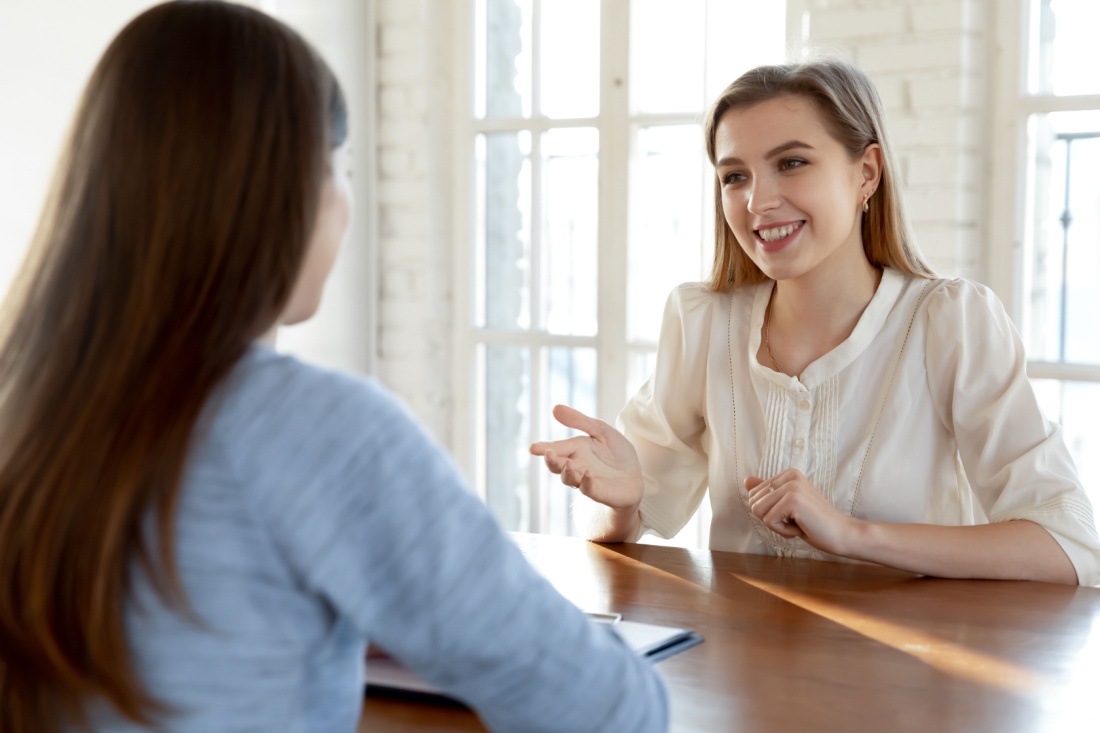 Two women sit across from each other, one is smiling and greeting the other warmly.