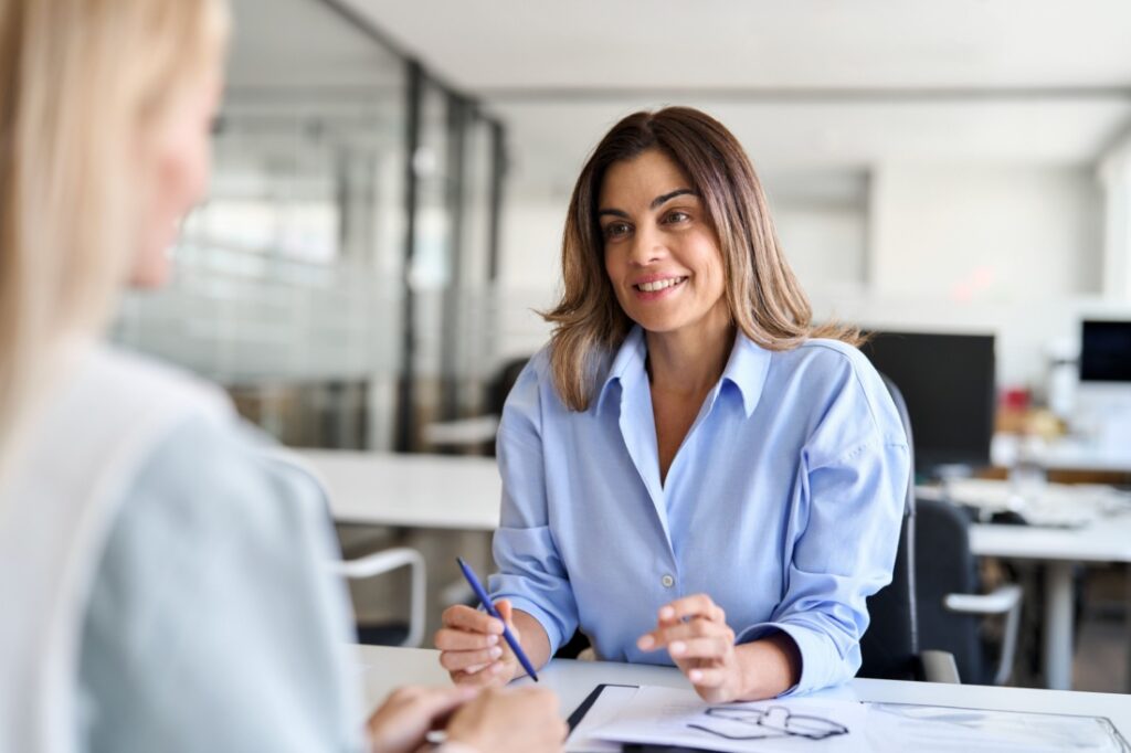 A professionally-dressed woman sits across from another woman, taking notes during a job interview.