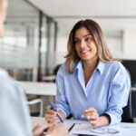 A professionally-dressed woman sits across from another woman, taking notes during a job interview.