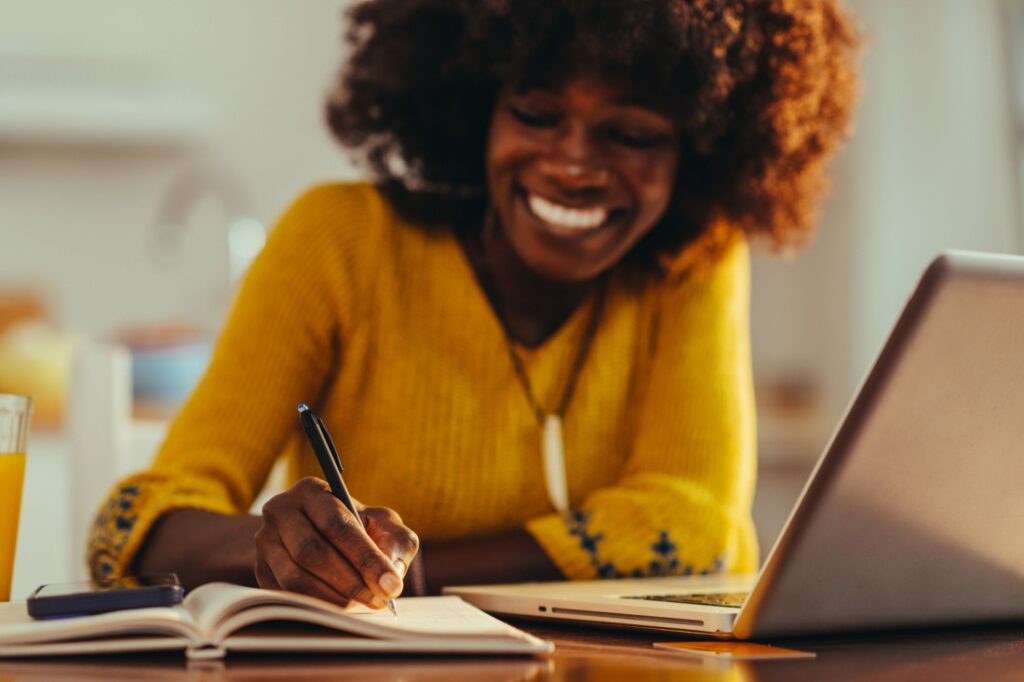 Selective focus on the businesswoman's hand, writing down tasks and notes in an agenda while working remotely from home. A happy interracial female entrepreneur is taking notes in the agenda.