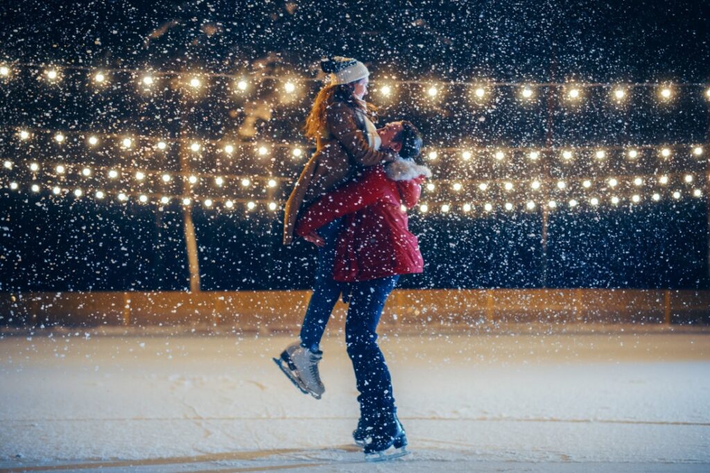 A snowy night. A man lifts up a woman, both are ice skating.