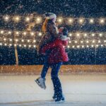 A snowy night. A man lifts up a woman, both are ice skating.