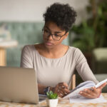 A Black woman looks at a laptop screen while writing in a notebook