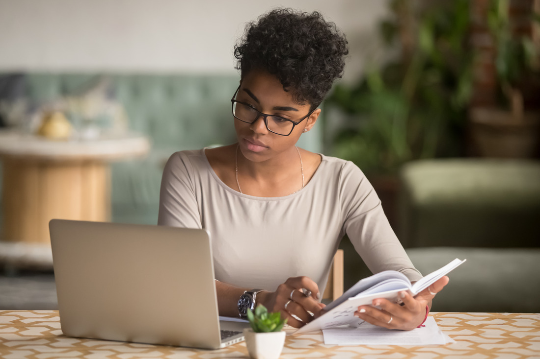 A Black woman looks at a laptop screen while writing in a notebook