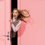 A woman peeks out from behind a pink door. a