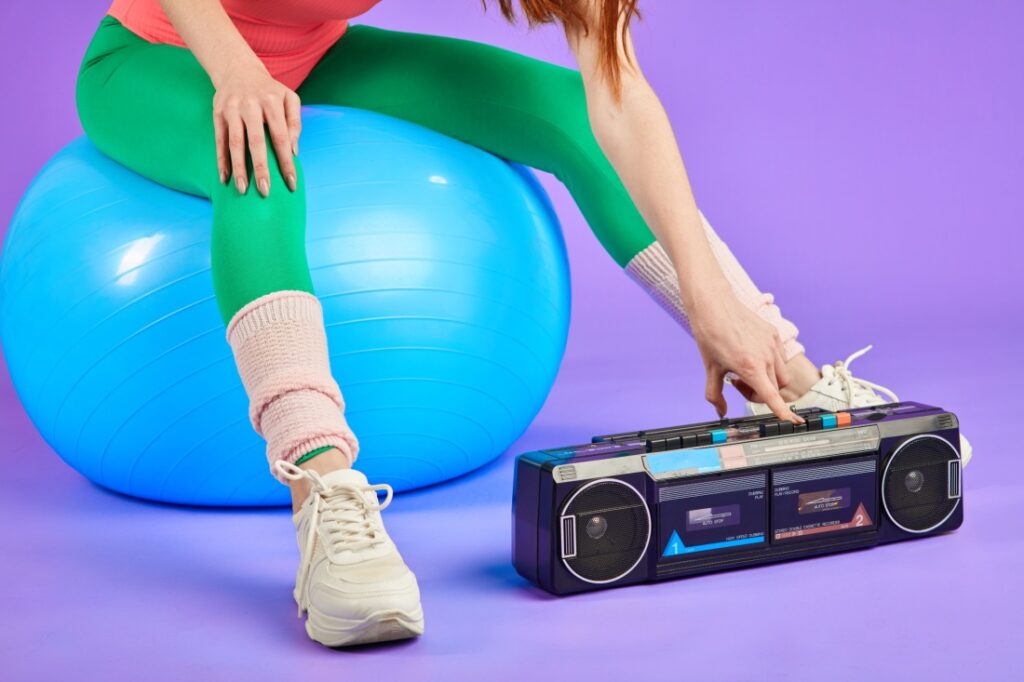 Cropped photo of a woman's legs in tights and leg warmers on an exercise ball, leaning over to turn on a stereo.