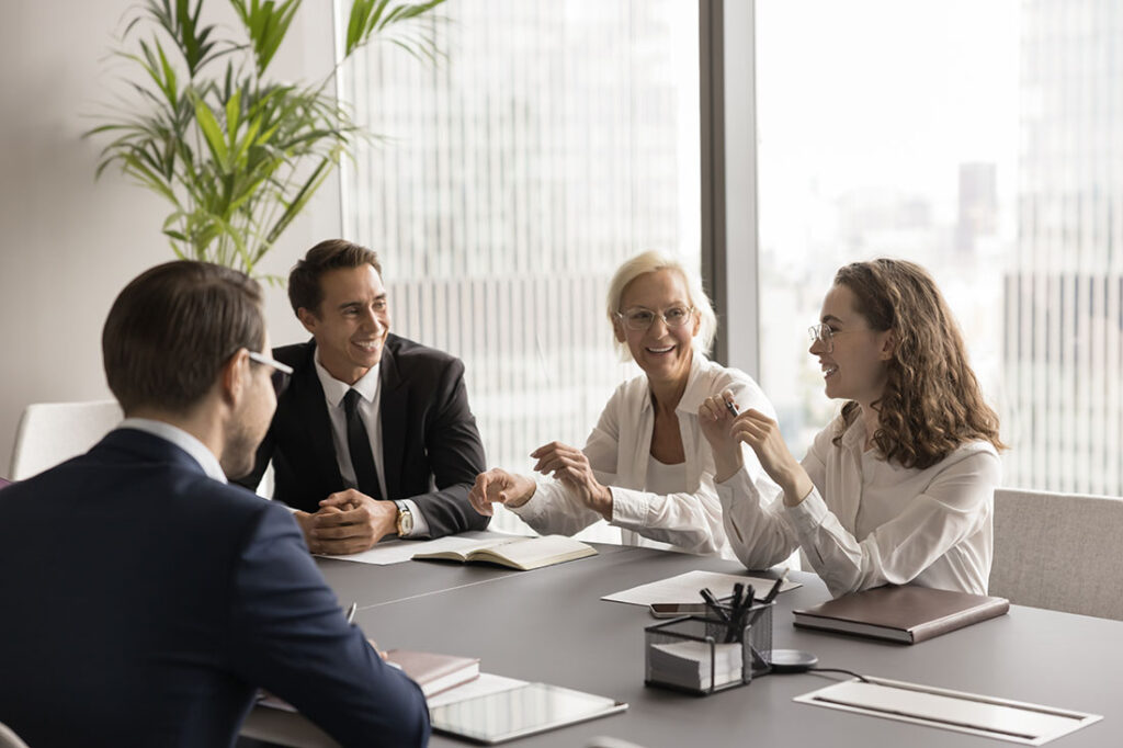 Positive elder and Gen Z coworkers laughing on corporate meeting, discussing creative ideas, brainstorming, having fun, sitting at conference table. Mature mentor, coach training employees