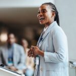 A confident woman gives a presentation, a blurred crowd behind her.