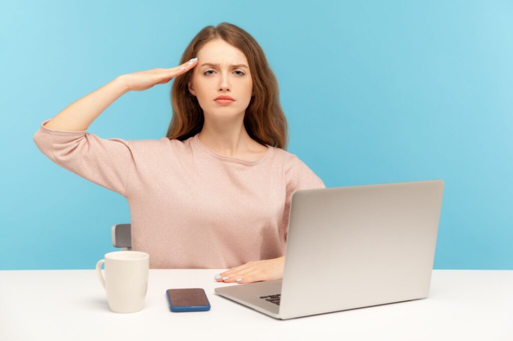 A woman salutes the camera. Her laptop, phone and coffee mug are in front of her.