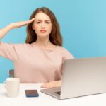 A woman salutes the camera. Her laptop, phone and coffee mug are in front of her.